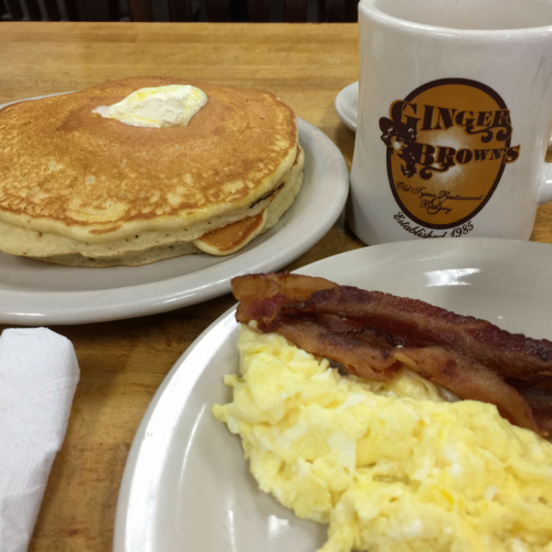 A plate with pancakes topped with butter, scrambled eggs, crispy bacon, and a coffee mug from Ginger Brown's restaurant.