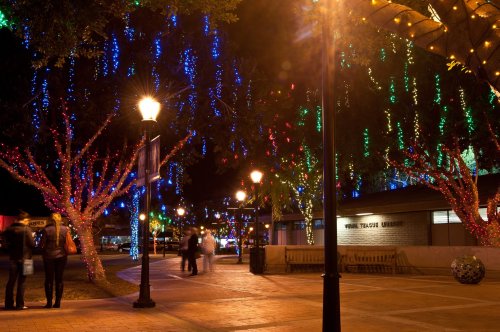 Colorful holiday lights adorn trees in a festive nighttime scene, with people walking and lampposts illuminating the area.