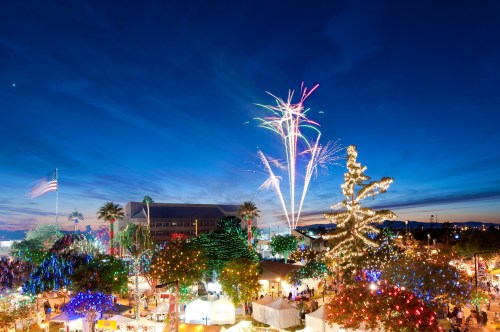 Colorful fireworks illuminate the night sky over a festive scene with decorated trees and lights, set against a blue backdrop.
