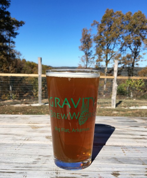 A pint of amber beer with a logo sits on a table, surrounded by trees and a clear blue sky in the background.