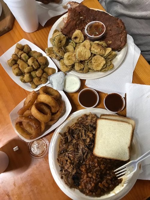 A table spread with barbecue meat, baked beans, fried okra, onion rings, and pickles, served with dipping sauces.