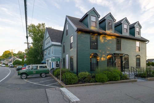 A green multi-story house with a vintage truck parked nearby, set on a quiet street with trees and a clear sky.