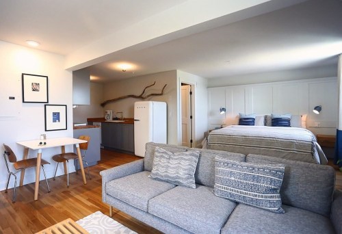 A cozy studio apartment featuring a bed, sofa, dining table, and kitchenette with modern decor and wooden accents.