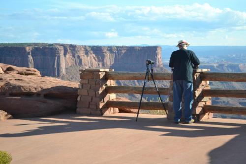 A person stands at a scenic overlook, photographing a vast canyon landscape with a tripod.