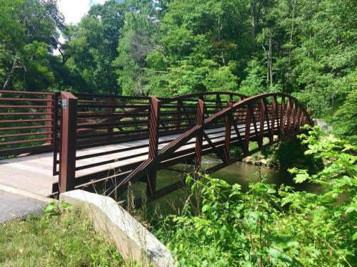 A curved metal bridge spans a small stream, surrounded by lush green trees and foliage.