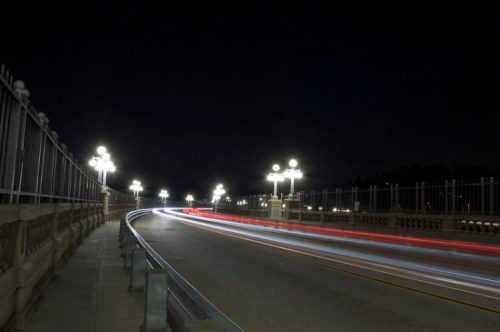 A nighttime view of a bridge with glowing street lamps and light trails from passing vehicles.