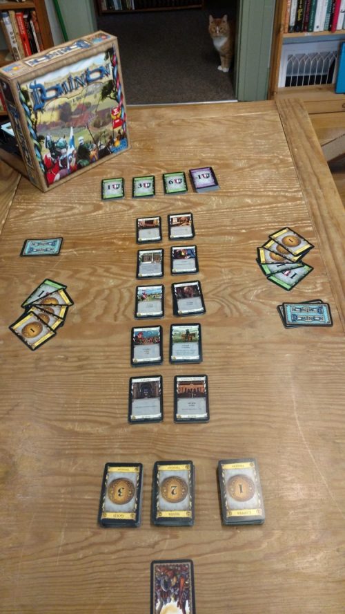 A tabletop game setup with cards arranged on a wooden table, a game box in the background, and a cat peeking in.