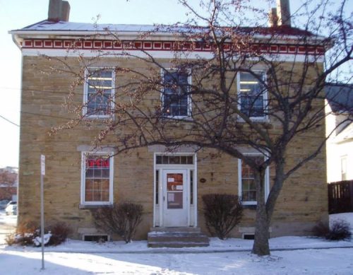A two-story stone building with a red trim, snow on the ground, and a tree in front. A sign in the window says "OPEN."