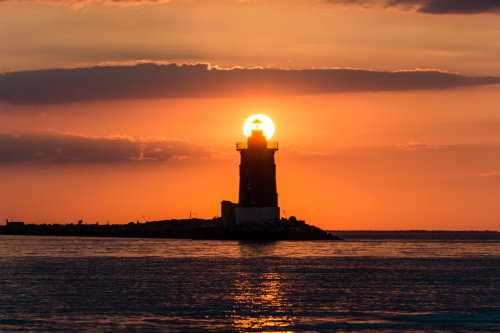 A silhouette of a lighthouse at sunset, with the sun glowing behind it, casting a warm orange hue over the water.