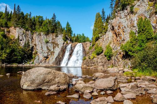 A serene waterfall cascades over rocky cliffs, surrounded by lush greenery and a calm river in a sunny landscape.