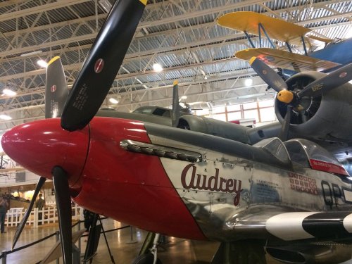 Close-up of a vintage aircraft with a red nose and the name "Audrey" painted on the side, displayed indoors.