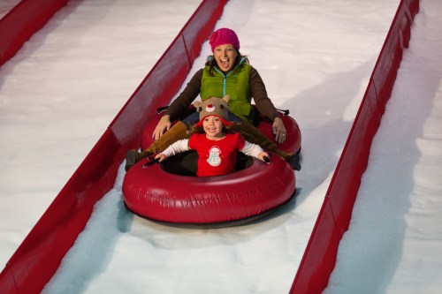 A woman and a child joyfully slide down a snowy slope on red inner tubes, both wearing winter hats.