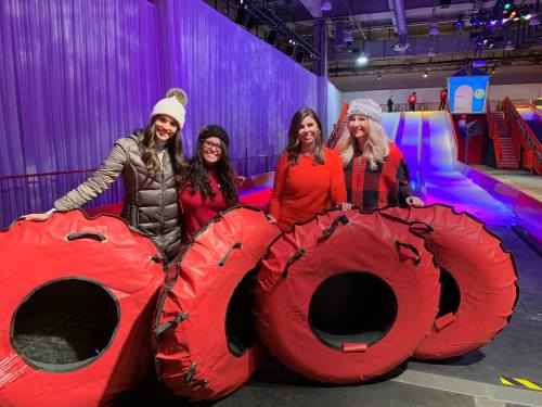 Four women pose with red snow tubes in a festive indoor winter setting, with colorful slides in the background.