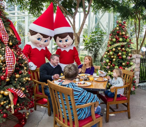 A family enjoys a festive meal surrounded by Christmas decorations and two large elf mascots.