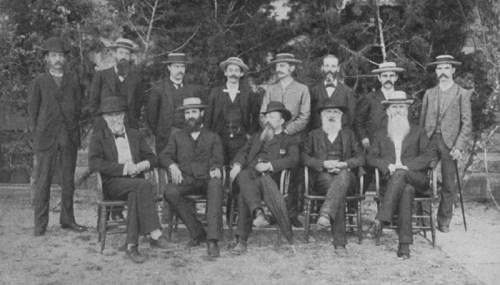 A historic black-and-white photo of twelve men in formal attire, seated and standing outdoors, with trees in the background.