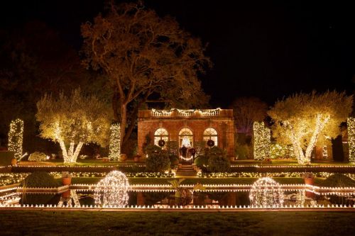 A beautifully lit garden at night, featuring a stone building, fountains, and trees adorned with twinkling lights.