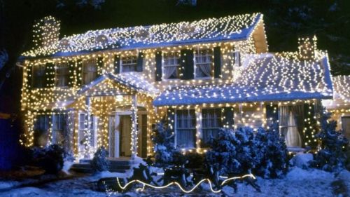 A festive house covered in bright white Christmas lights, surrounded by snow and trees, creating a cheerful holiday scene.