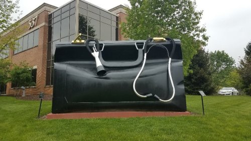A large sculpture of a black briefcase with a stethoscope, set in a grassy area near a brick building.