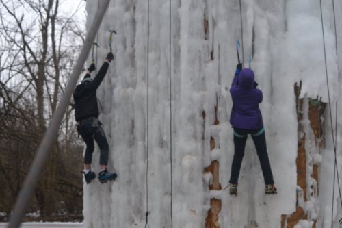 Two climbers ascend an icy wall, using ice axes and wearing winter gear, surrounded by bare trees.