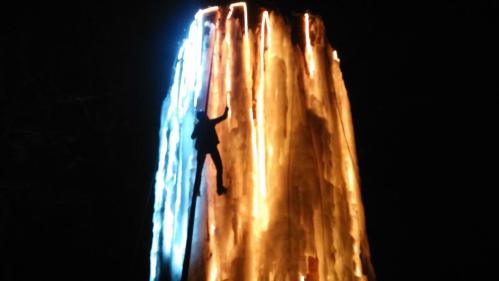 A climber ascends a brightly lit, ice-covered tower at night, illuminated in blue and orange hues.