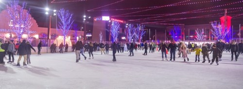 A lively ice skating rink at night, filled with people skating under colorful lights and festive decorations.