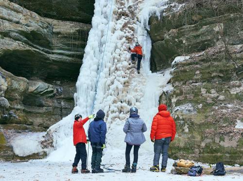 A climber ascends a frozen waterfall while four people observe and assist from the ground in a winter landscape.