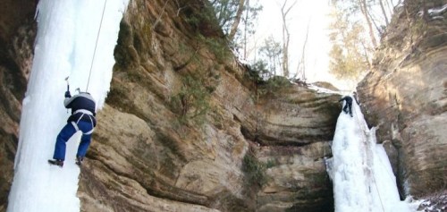 Two climbers ascend icy rock formations in a wooded canyon during winter.