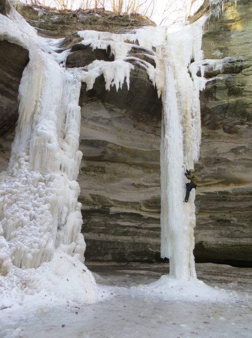 A climber ascends a frozen waterfall surrounded by icy rock formations in a winter landscape.