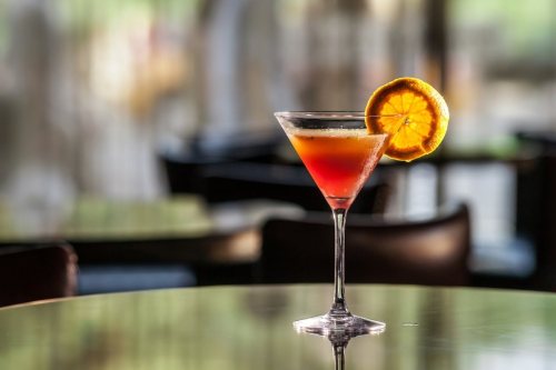 A colorful cocktail in a martini glass, garnished with a slice of orange, sits on a reflective table.