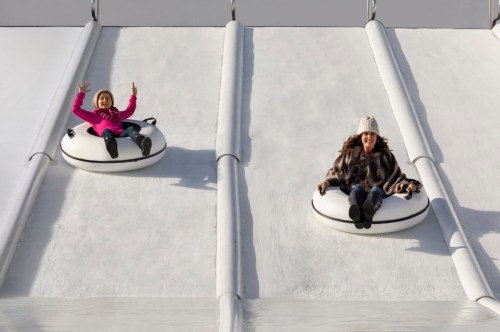 Two women on snow tubes sliding down a snowy slope, one waving and smiling, wearing winter clothing.