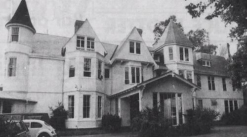A large, historic mansion with multiple gables and a front porch, surrounded by trees and greenery.