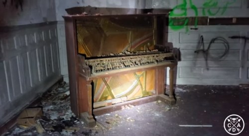 An old, dusty piano sits in a dimly lit, abandoned room, surrounded by debris and graffiti on the walls.