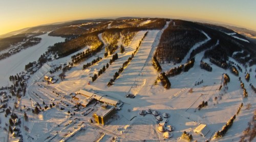 Aerial view of a snowy landscape with a ski resort, trees, and hills under a clear sky during sunset.