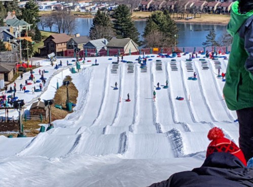 A snowy hill with people sledding down, surrounded by trees and houses, near a lake in the background.