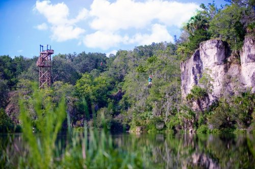 A person zips across a lake on a zip line, with a wooden tower and lush greenery in the background.