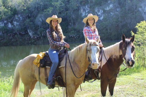 Two women in cowboy hats sit on horses by a serene lake, surrounded by lush greenery and rocky cliffs.