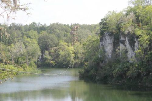 A scenic view of a lake surrounded by cliffs and trees, featuring a zip line tower in the distance.