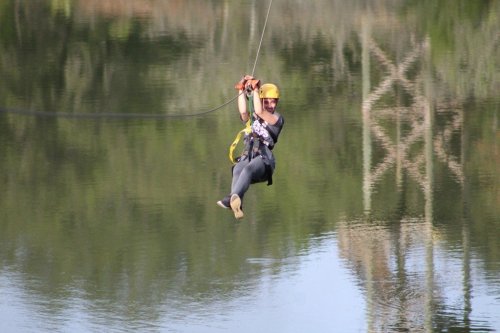 A person in a yellow helmet zip-lining over a calm body of water, surrounded by greenery.