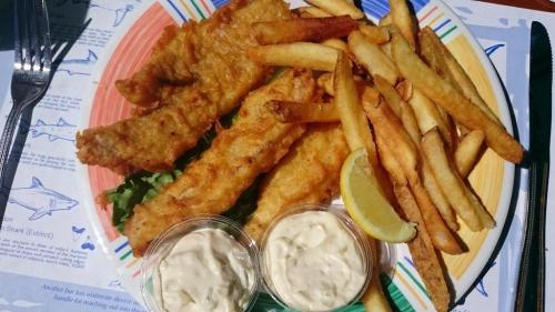 A colorful plate with crispy fish fillets, golden fries, a lemon wedge, and two cups of dipping sauce on a patterned placemat.