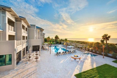 A beachfront hotel with a pool area, palm trees, and a sunset over the ocean in the background.