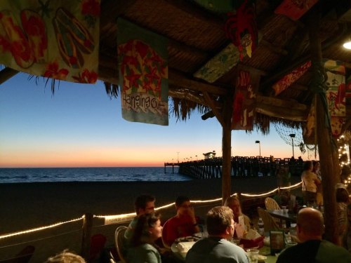 A beachside restaurant at sunset, with colorful banners and a pier in the distance, creating a warm, inviting atmosphere.