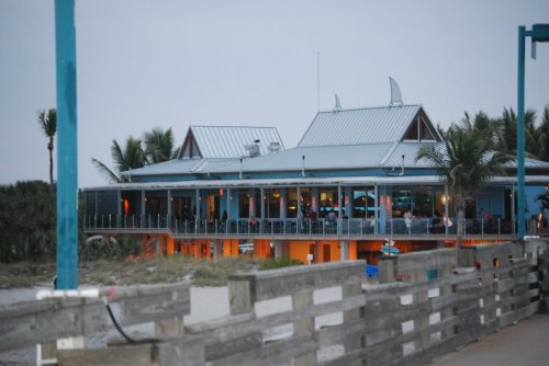 A beachside restaurant with a blue exterior and palm trees, viewed from a wooden pier at dusk.