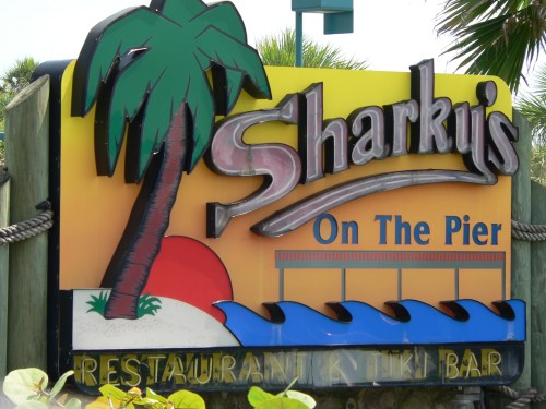 Colorful sign for Sharky's On The Pier, featuring a palm tree, sunset, and waves, indicating a restaurant and tiki bar.
