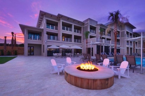A modern hotel exterior at sunset, featuring a fire pit, lounge chairs, and palm trees by the pool.