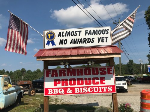 Sign for a BBQ and produce stand featuring flags, stating "Almost Famous No Awards Yet" and promoting BBQ and biscuits.