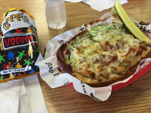 A sandwich topped with melted cheese and lettuce, served with a pickle and a bag of Voodoo chips on a wooden table.