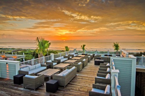A scenic view of a beachfront deck with lounge seating, overlooking the ocean at sunset with colorful clouds.
