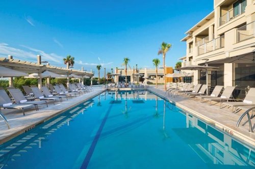 A sunny pool area with lounge chairs, palm trees, and modern buildings in the background. Clear blue sky above.