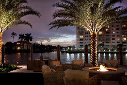 A cozy outdoor seating area with a fire pit, palm trees, and a waterfront view at dusk.