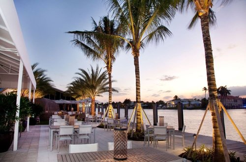 Outdoor dining area by the water, featuring palm trees, modern furniture, and a sunset backdrop.